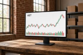 VeChain (VET) Nedir? Hayabusa, VTHO ve Sabit Arz Modeli 34 Tuğla duvarlı depo ofisinde ahşap masadaki açık temalı monitörde VET mum grafiği ve hacim çubukları, VeChain için temsili görsel